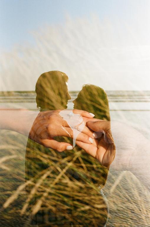 A double exposure shows a couple embracing and a woman's hand with an engagement ring over a field of tall grass with the ocean in the background.