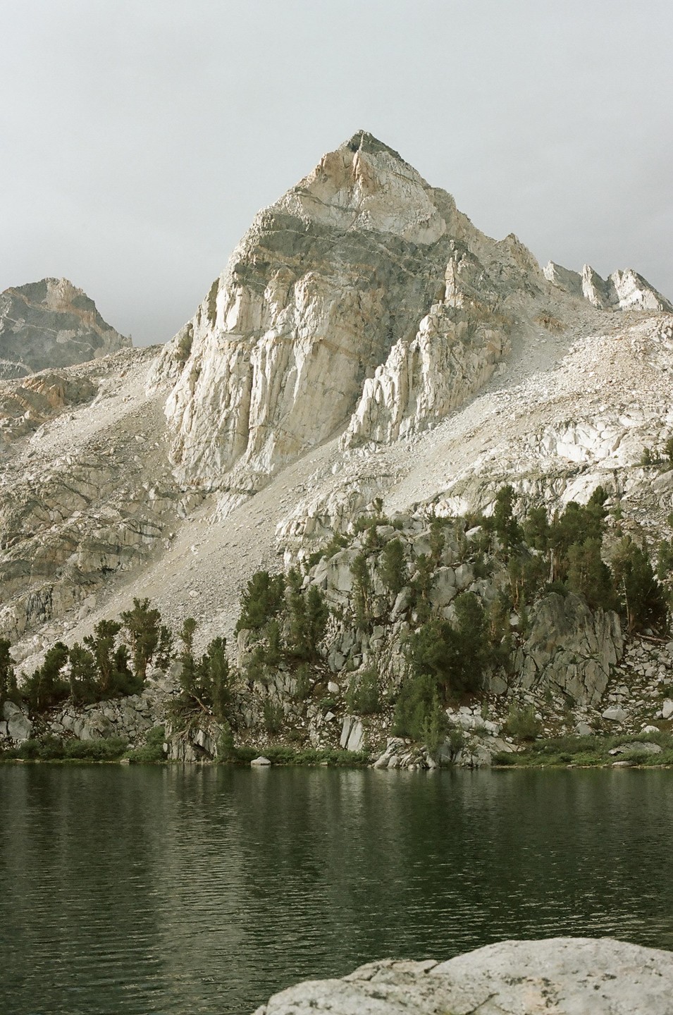 A large mountain rises above a lake with trees and rocks on its shore.