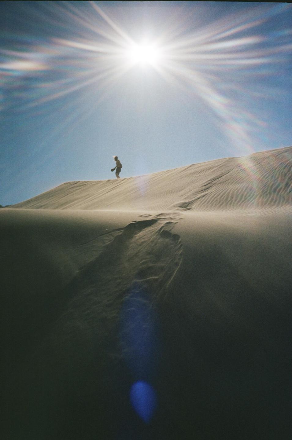 A person walks along the ridge of a sand dune under a bright sun.