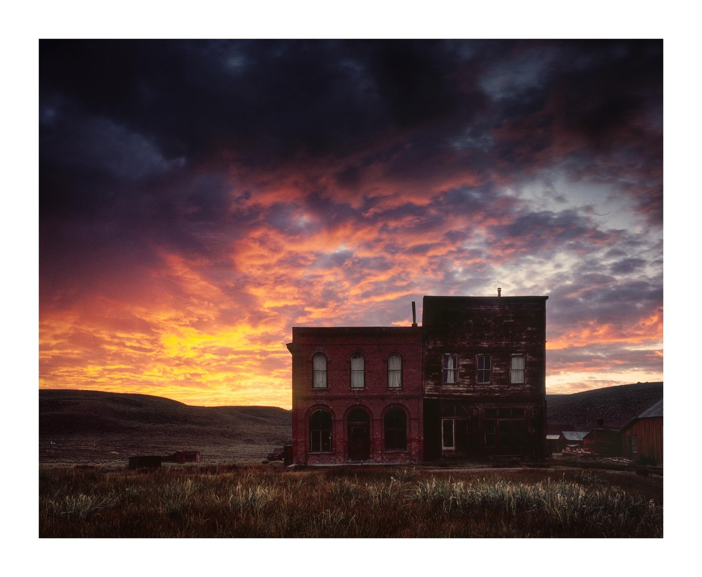 Two old buildings stand in a field under a dramatic sunset sky.