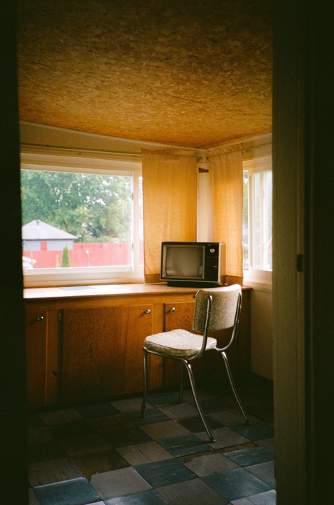 A vintage television sits on a wooden cabinet next to a window with yellow curtains, with a chair in the foreground and a checkered floor.