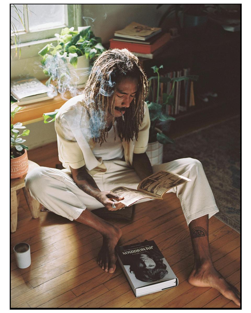 A man with dreadlocks sits on a wooden floor, smoking and reading a book, with plants and books in the background.
