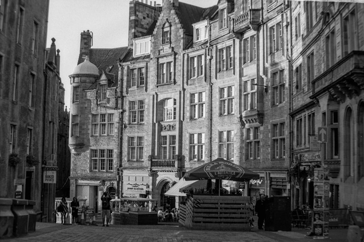 A street in Edinburgh is lined with tall, ornate buildings, with people walking and sitting at outdoor cafes.