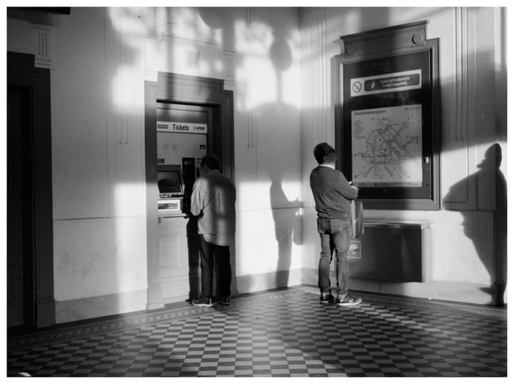 Two men stand in a room with a checkered floor, one at a ticket machine and the other looking at a map.