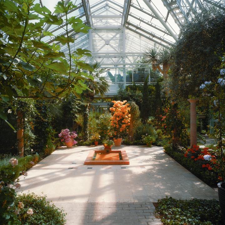 A lush conservatory features a central tiled patio with a small fountain, surrounded by diverse plants and flowers under a glass roof.