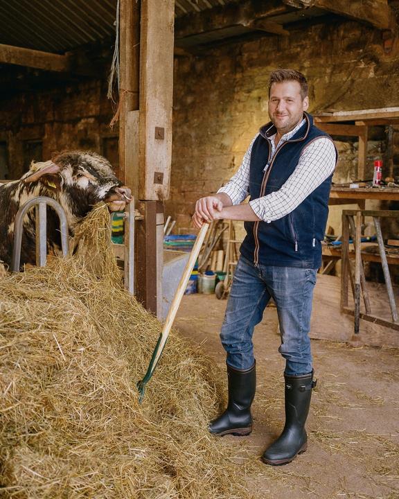 A man stands with a pitchfork next to a cow eating hay in a barn.