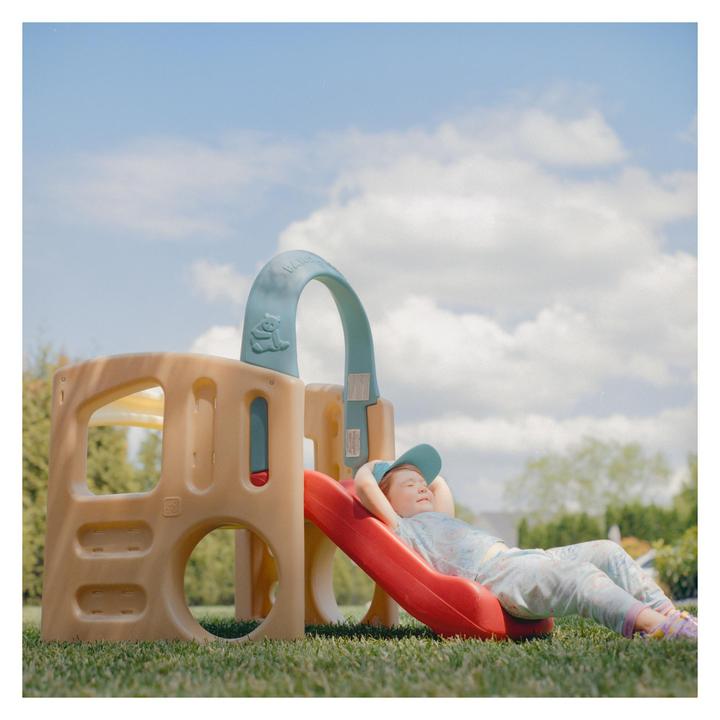 A child wearing a baseball cap is lying on a red slide in a grassy yard under a blue sky with clouds.