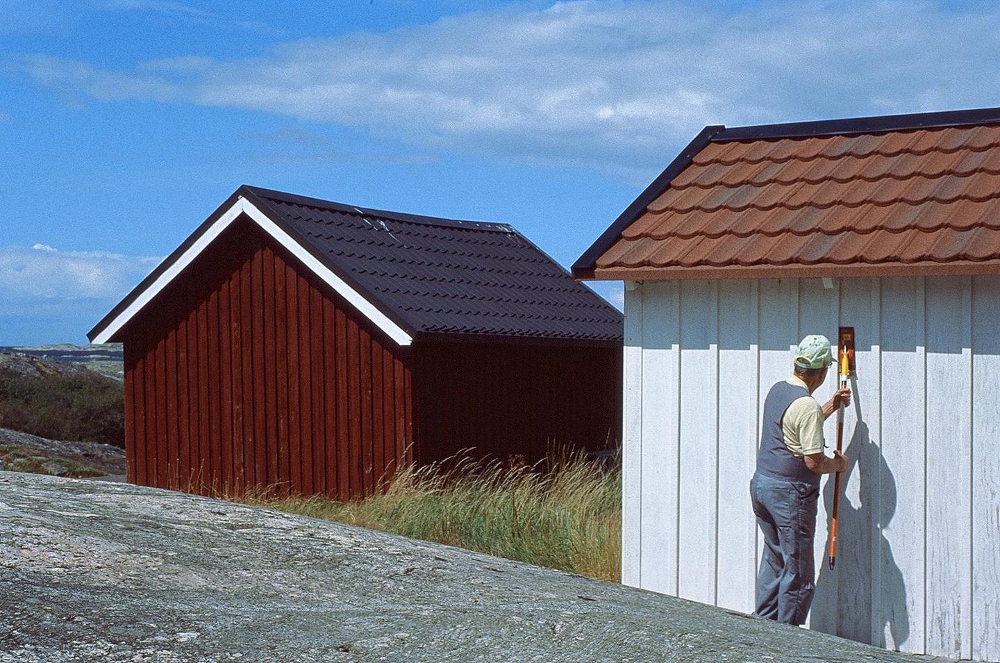 A man in a cap and overalls uses a tool to work on the side of a white building with a red roof, next to a red building with a black roof, under a blue sky.