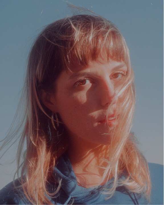 A woman with bangs and long hair looks directly at the viewer, with her hair blowing across her face.