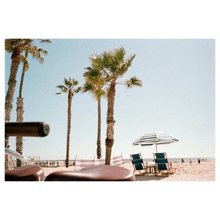 A beach scene features palm trees, a striped umbrella, and two beach chairs on the sand, with a bicycle seat in the foreground.