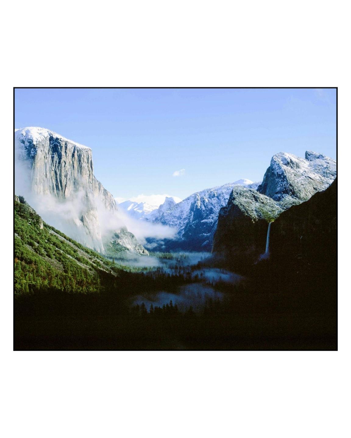 A valley with snow-capped mountains and a waterfall is covered in fog.