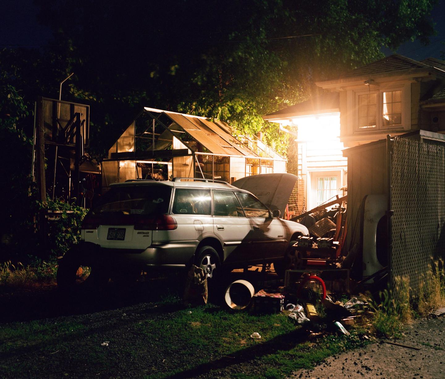 A car with its hood open is parked next to a greenhouse and a house at night.