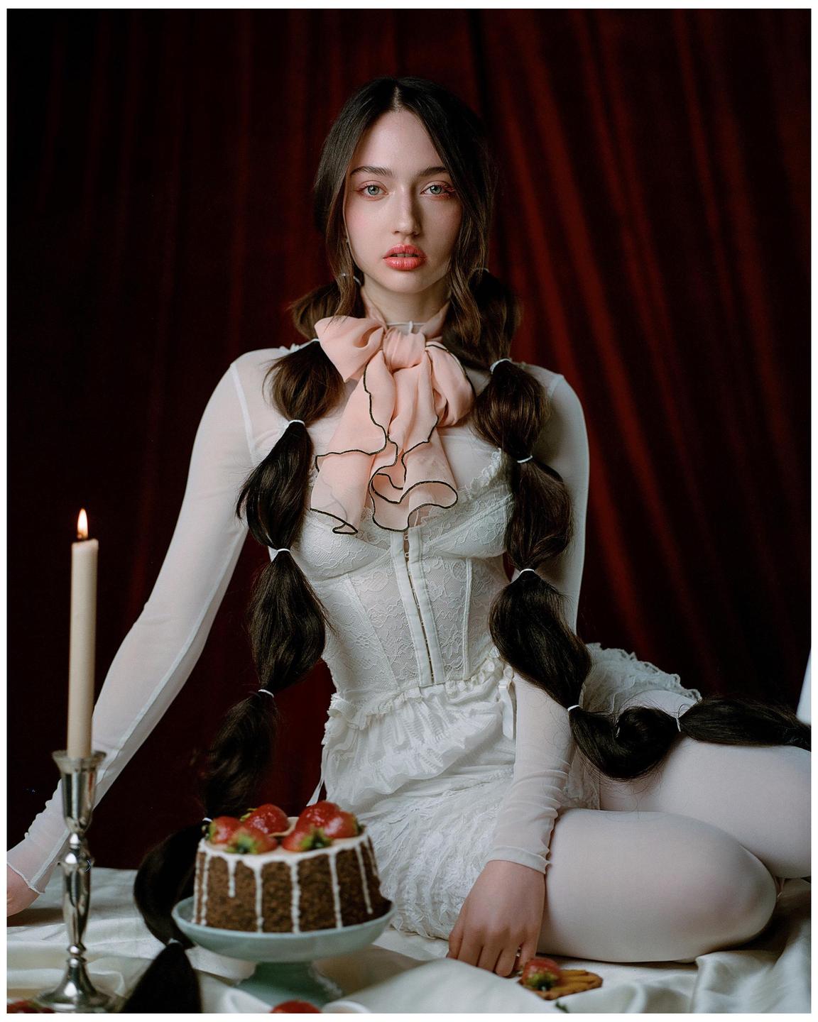 A woman with long braided hair and a large pink bow sits next to a chocolate cake with strawberries and a lit candle.