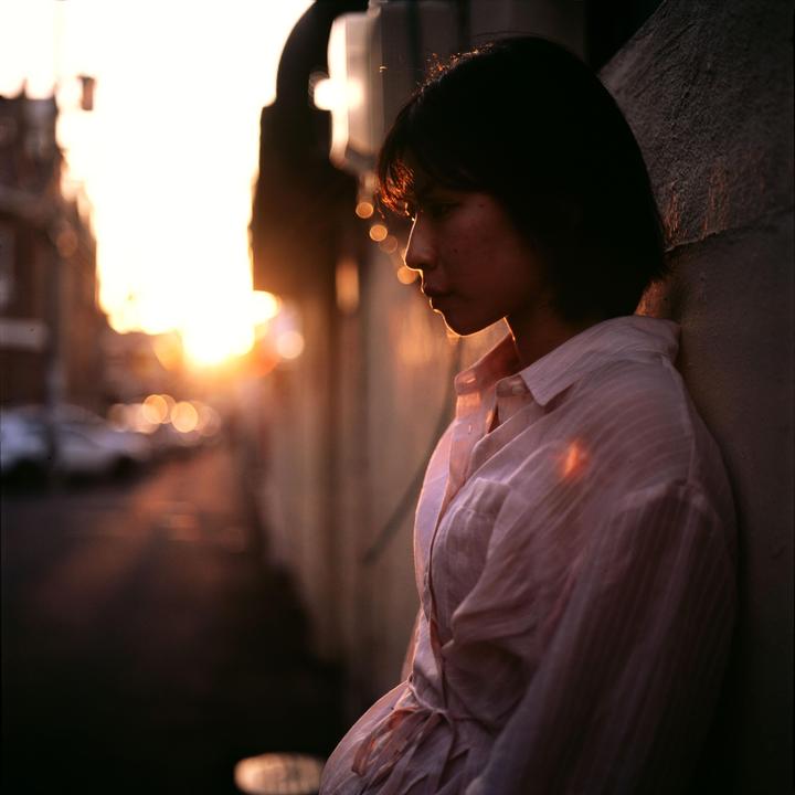 A woman stands in profile against a wall with a bright sunset in the background.