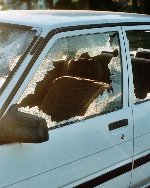 The side of a white car with shattered windows is illuminated by sunlight.