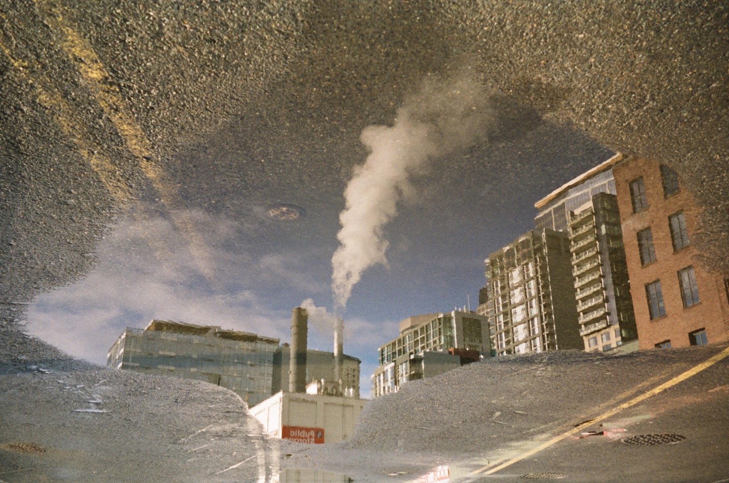 A puddle reflects a city street with buildings and a smoking chimney under a cloudy sky.