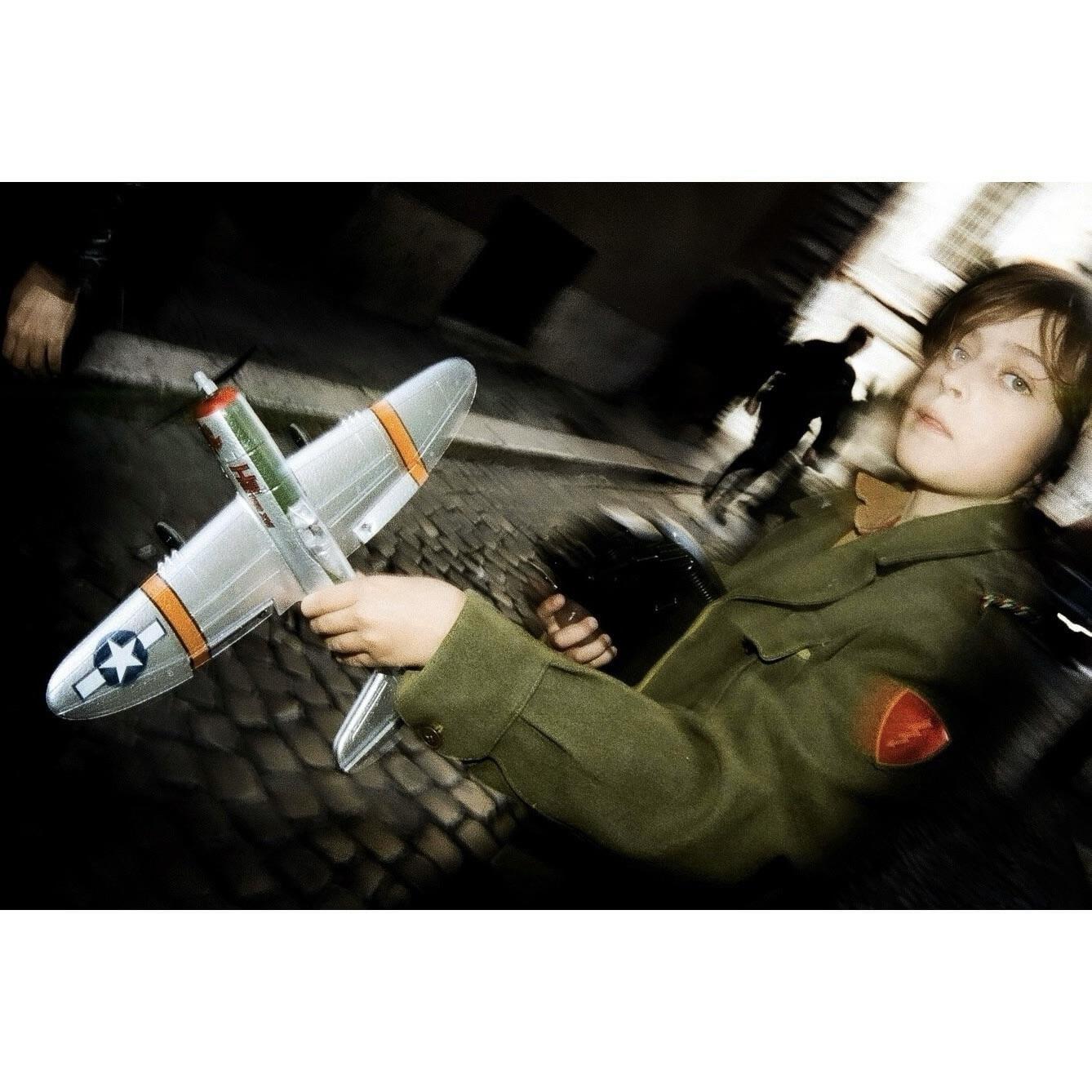 A child in a green jacket holds a model airplane while looking at the viewer, with a blurred street and a silhouetted figure in the background.