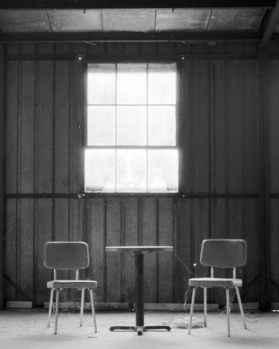 Two chairs and a table are centered in front of a large window in a room with corrugated metal walls.