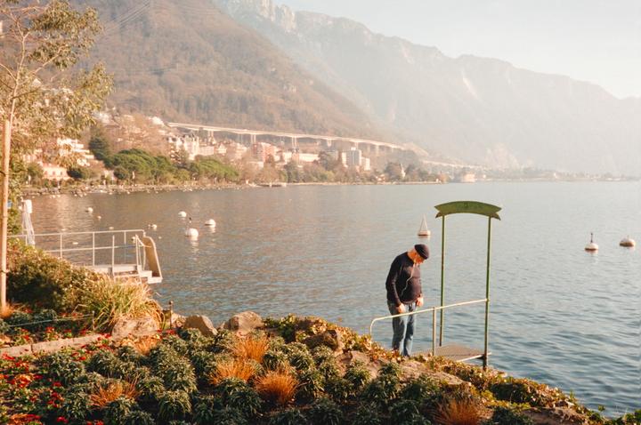 A man stands on a small dock by a lake with mountains and a town in the background.