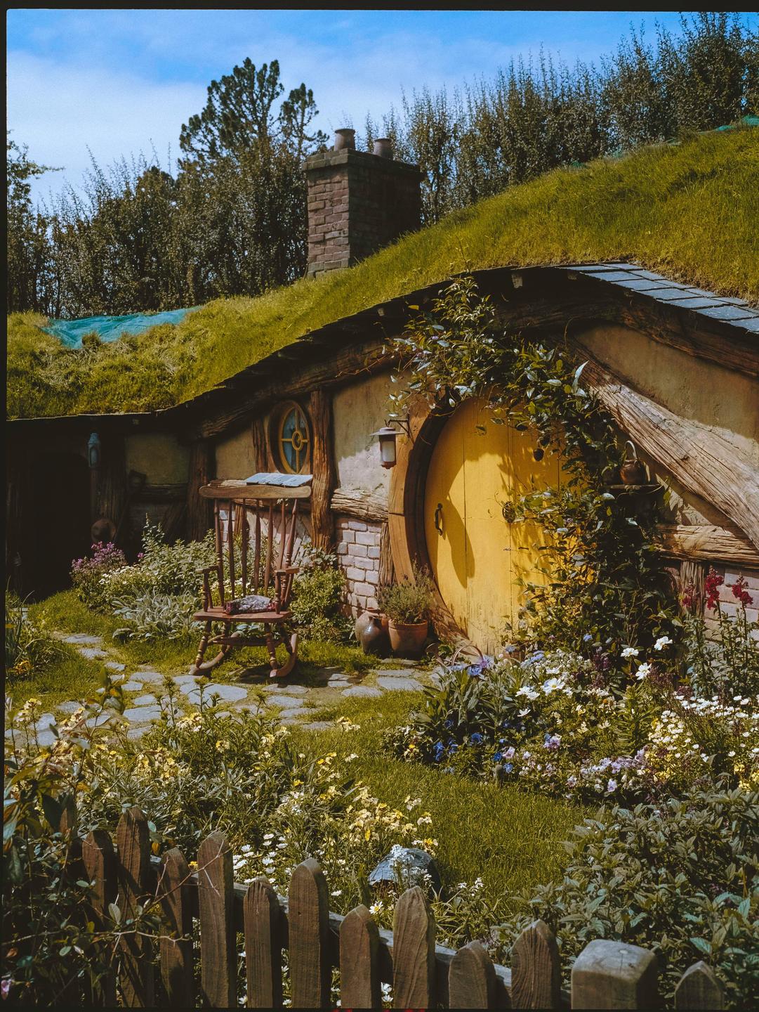 A hobbit hole with a round yellow door is nestled into a grassy hill, surrounded by a garden and a wooden fence.