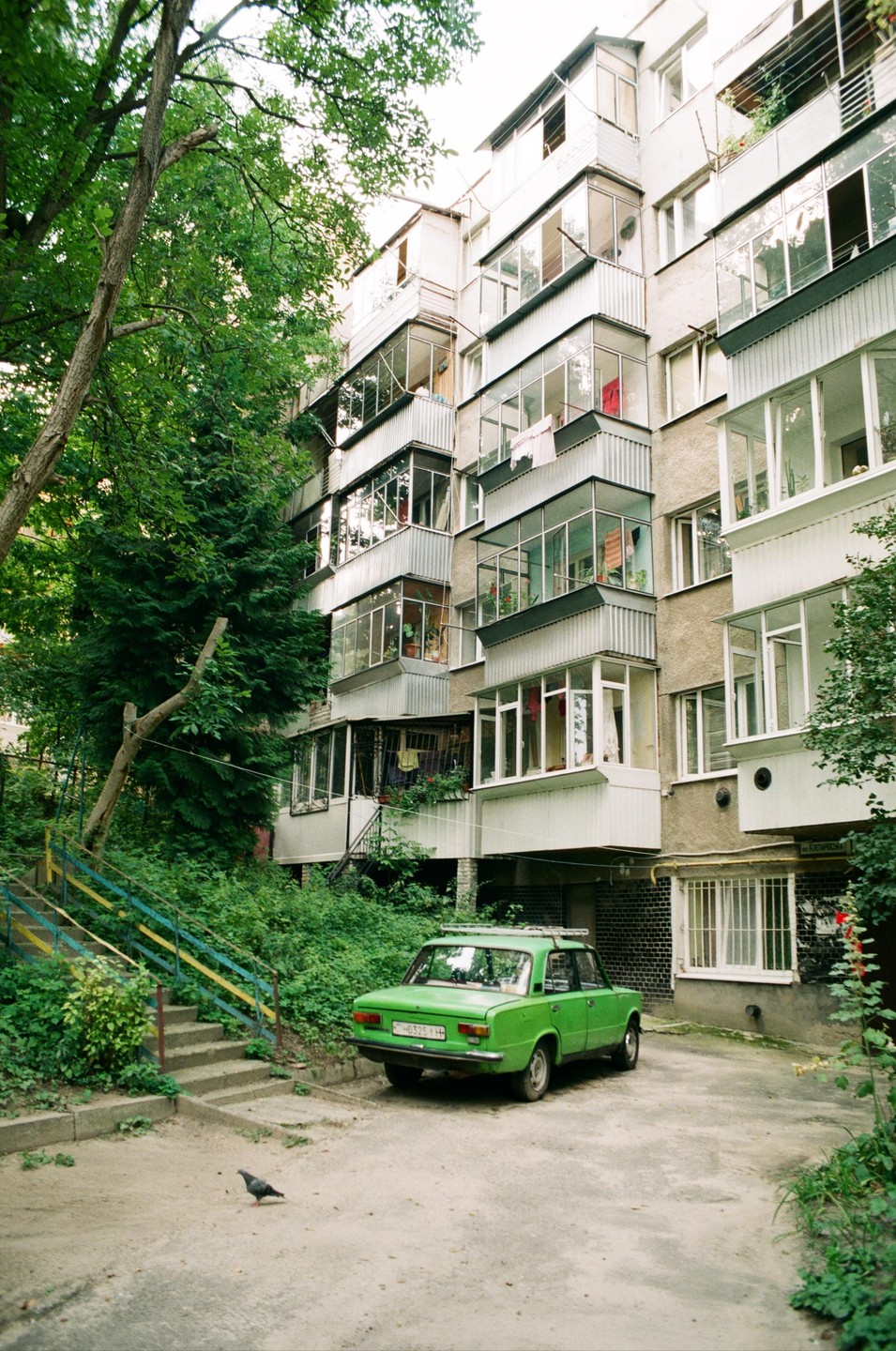 A green car is parked in front of a multi-story building with many balconies, next to a lush green hillside.