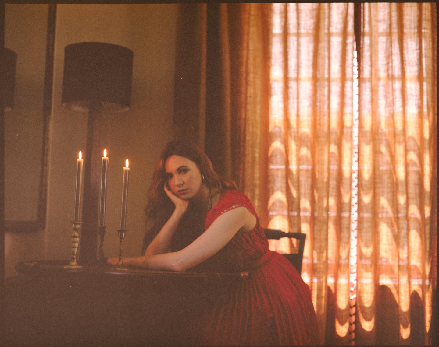 A woman in a red dress rests her head on her hand at a table with three lit candles.