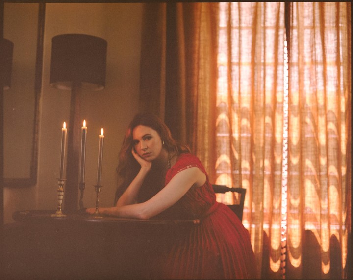 A woman in a red dress rests her head on her hand at a table with three lit candles.