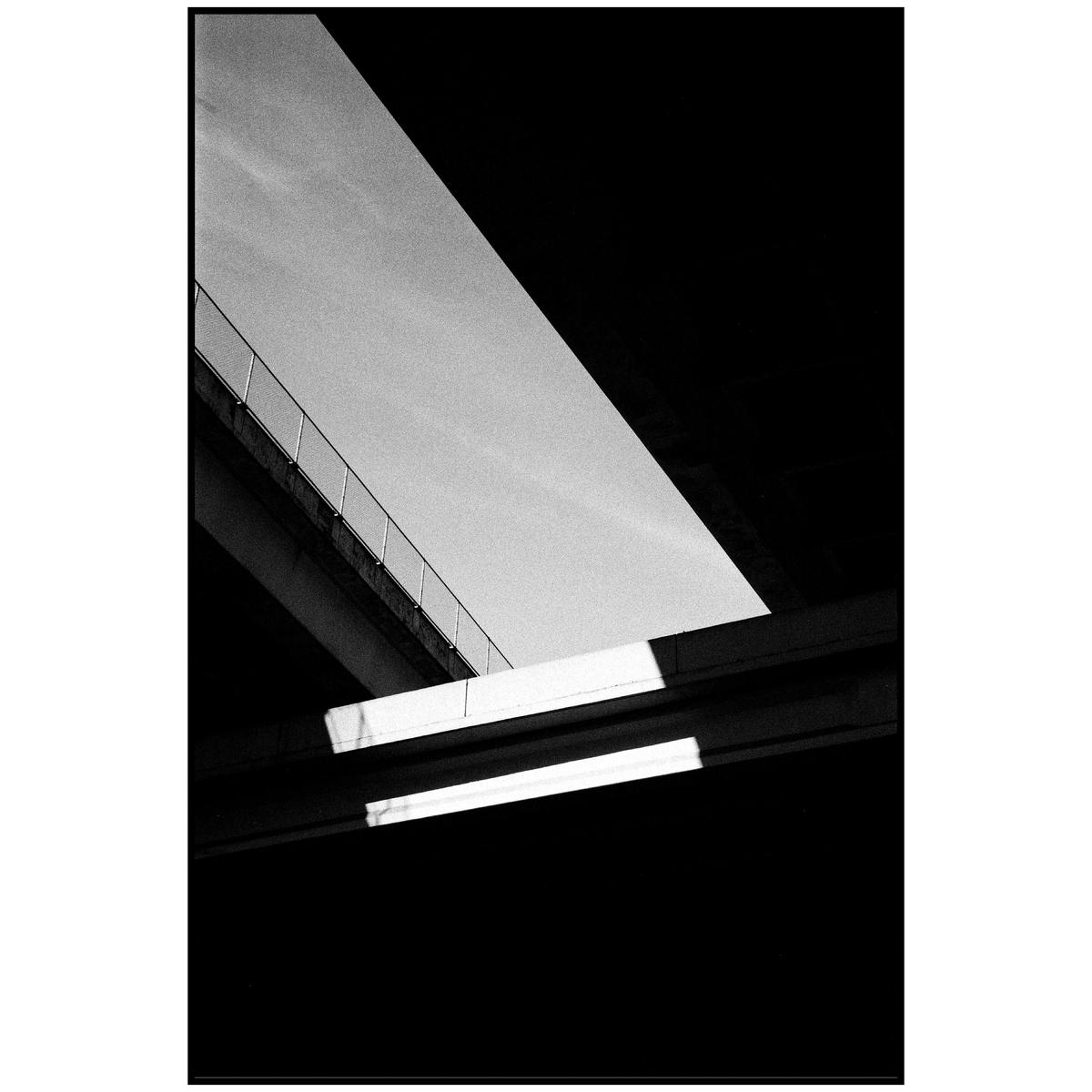 A black and white abstract image shows a diagonal concrete overpass with a railing, and two fluorescent lights below.