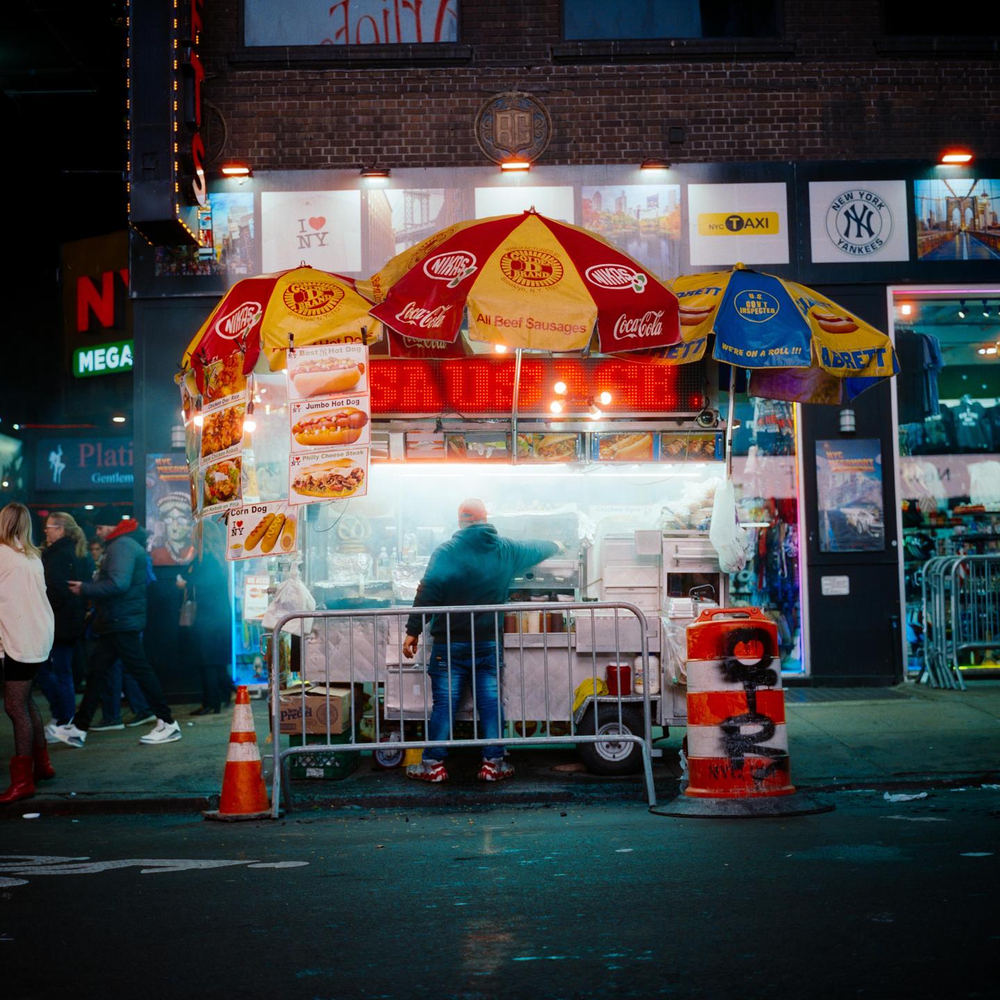 A man stands behind a brightly lit hot dog stand with red and yellow umbrellas on a city street at night.
