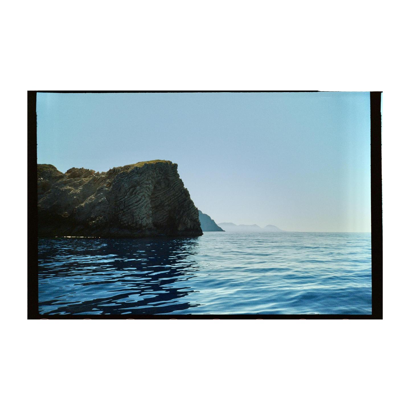 A large rock formation rises from the calm blue water under a clear sky.