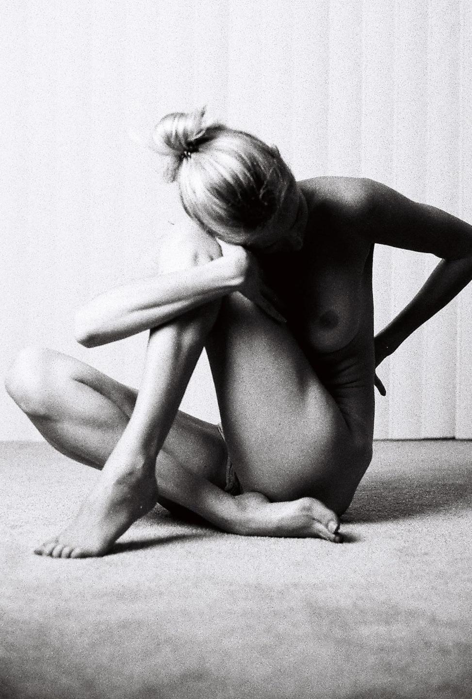 A woman with her hair in a bun sits on the floor with her head bowed and arms crossed over her knees.