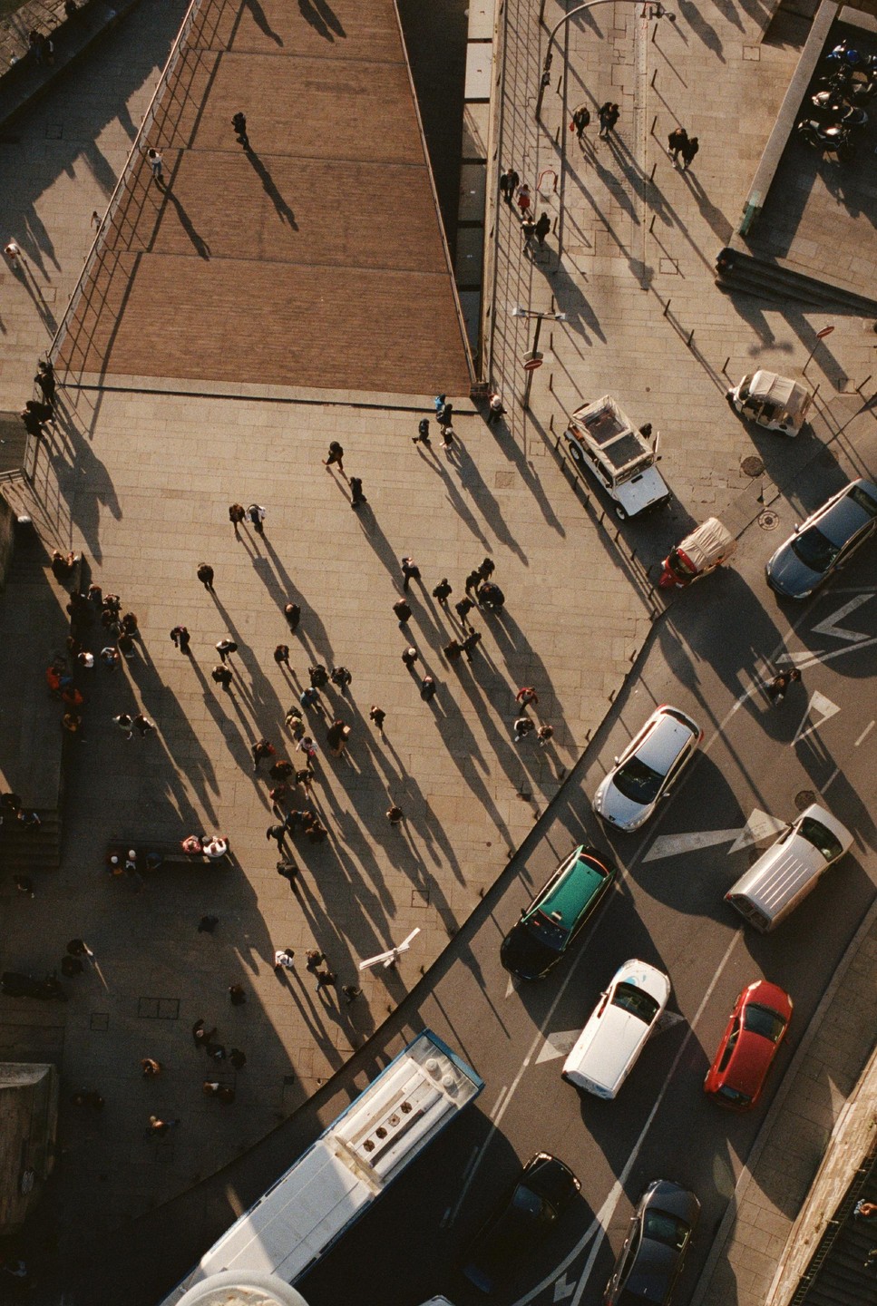 An aerial view shows people and vehicles on a city street with long shadows stretching across the pavement.