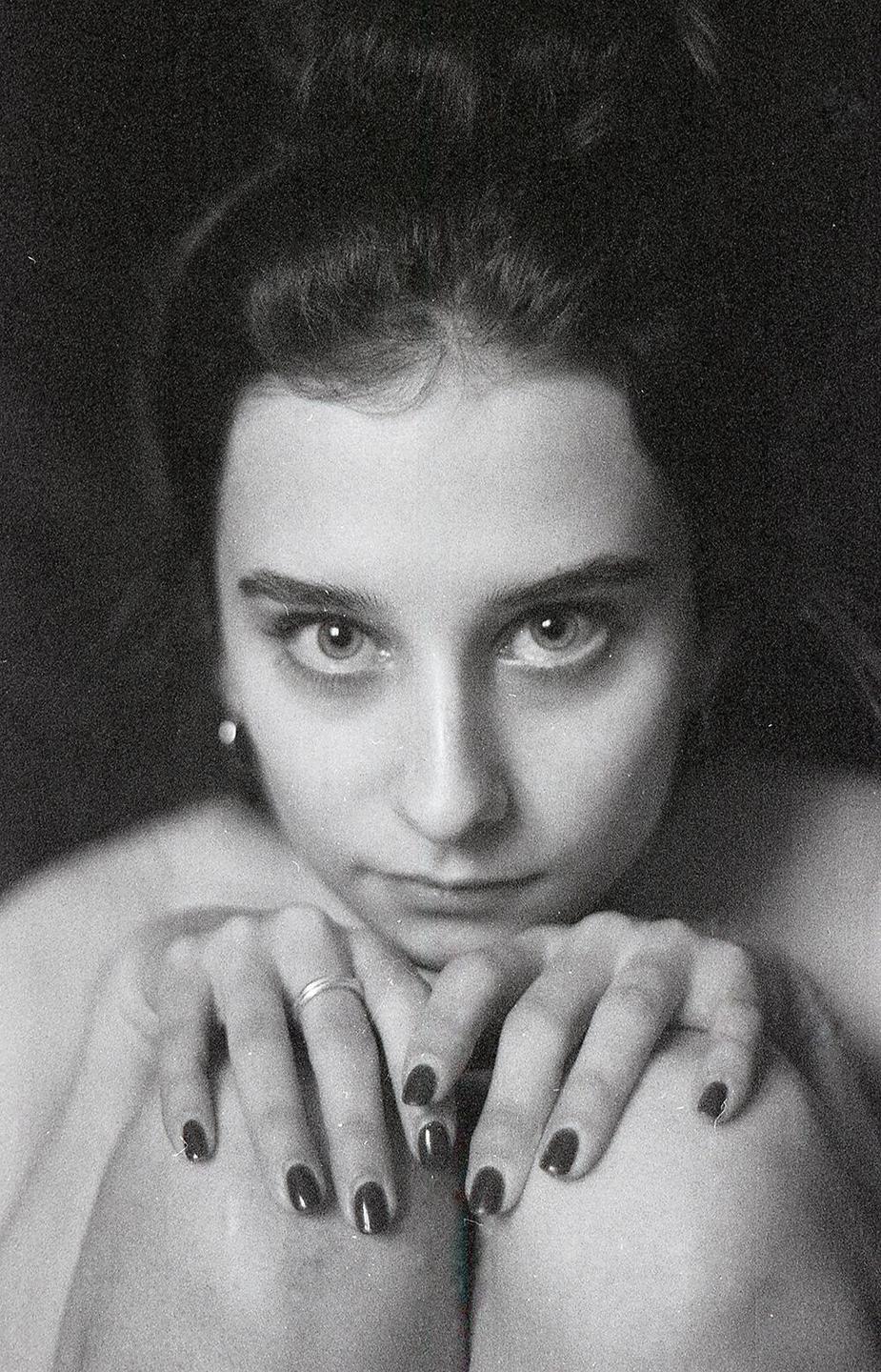 A woman with dark hair and dark nail polish looks directly at the viewer.
