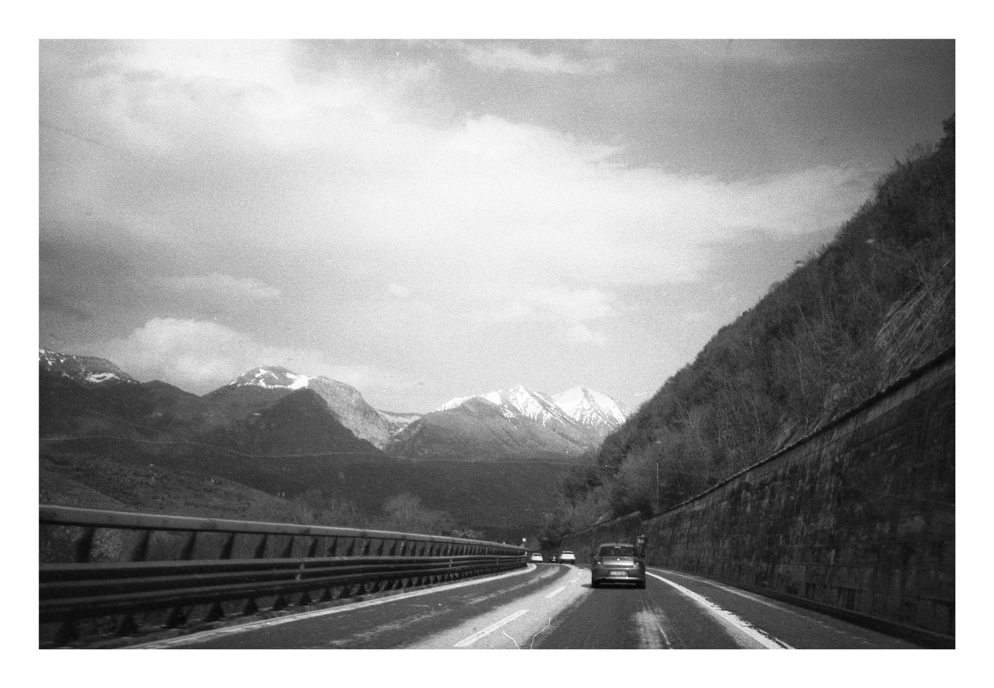 A car drives on a highway through a mountainous landscape with snow-capped peaks under a cloudy sky.