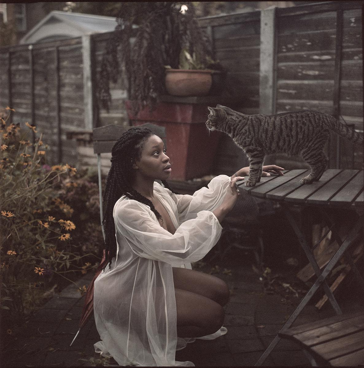 A woman in a sheer white robe crouches in a garden, looking up at a tabby cat standing on a wooden table.