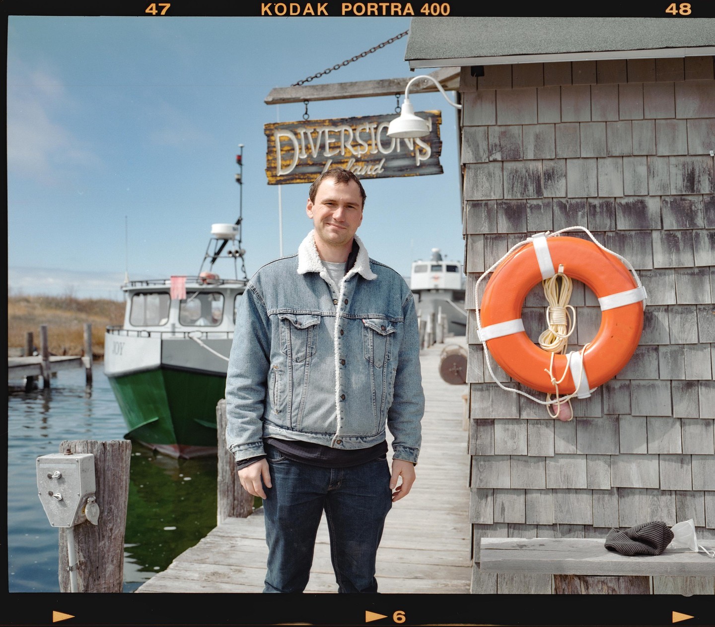 A man stands on a wooden dock next to a building with a life preserver hanging on it, with a boat in the water behind him.