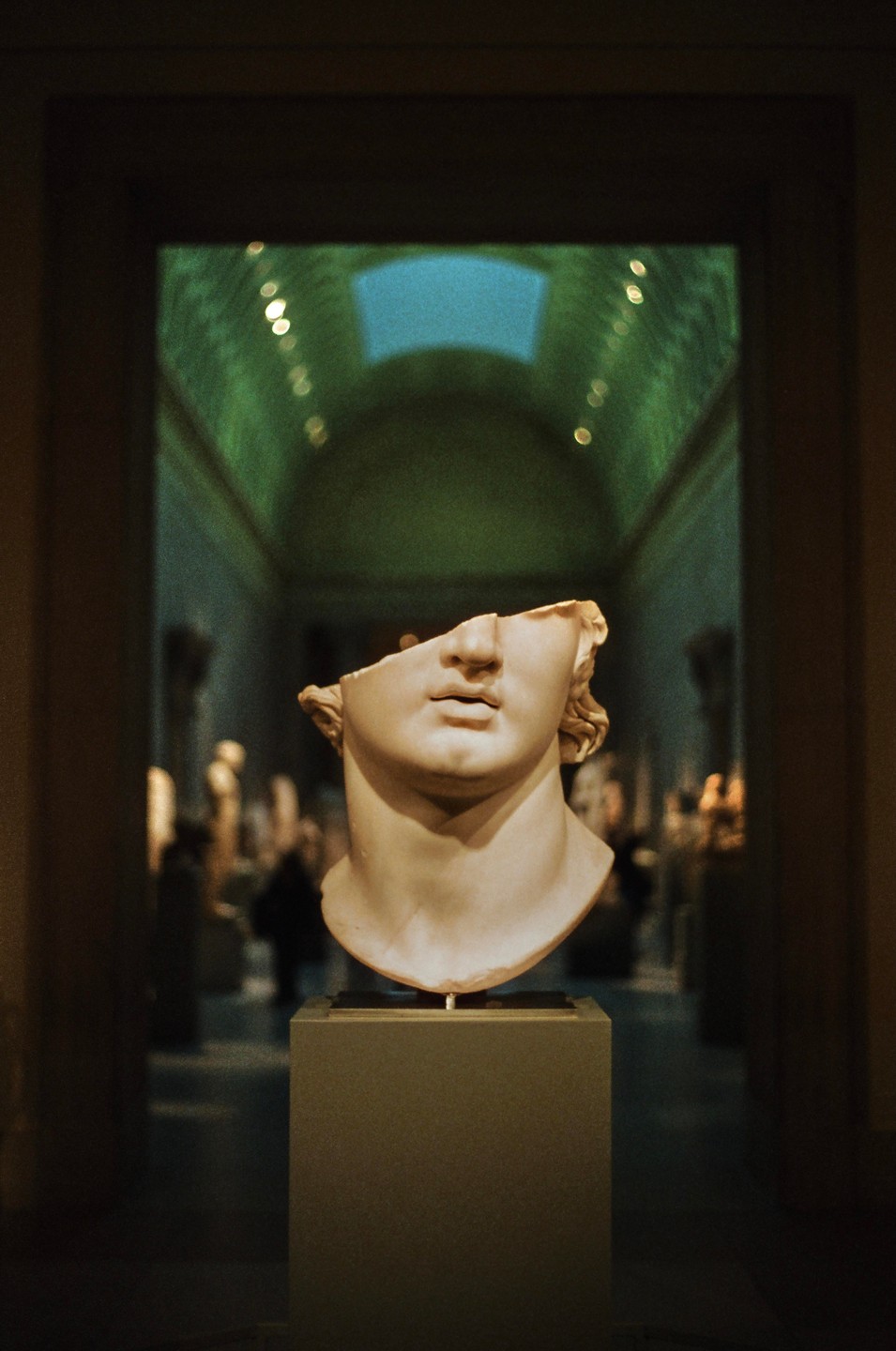 A broken marble bust of a man is displayed in a museum hall with a green ceiling and a skylight.