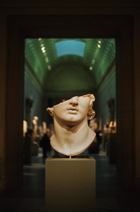A broken marble bust of a man is displayed in a museum hall with a green ceiling and a skylight.