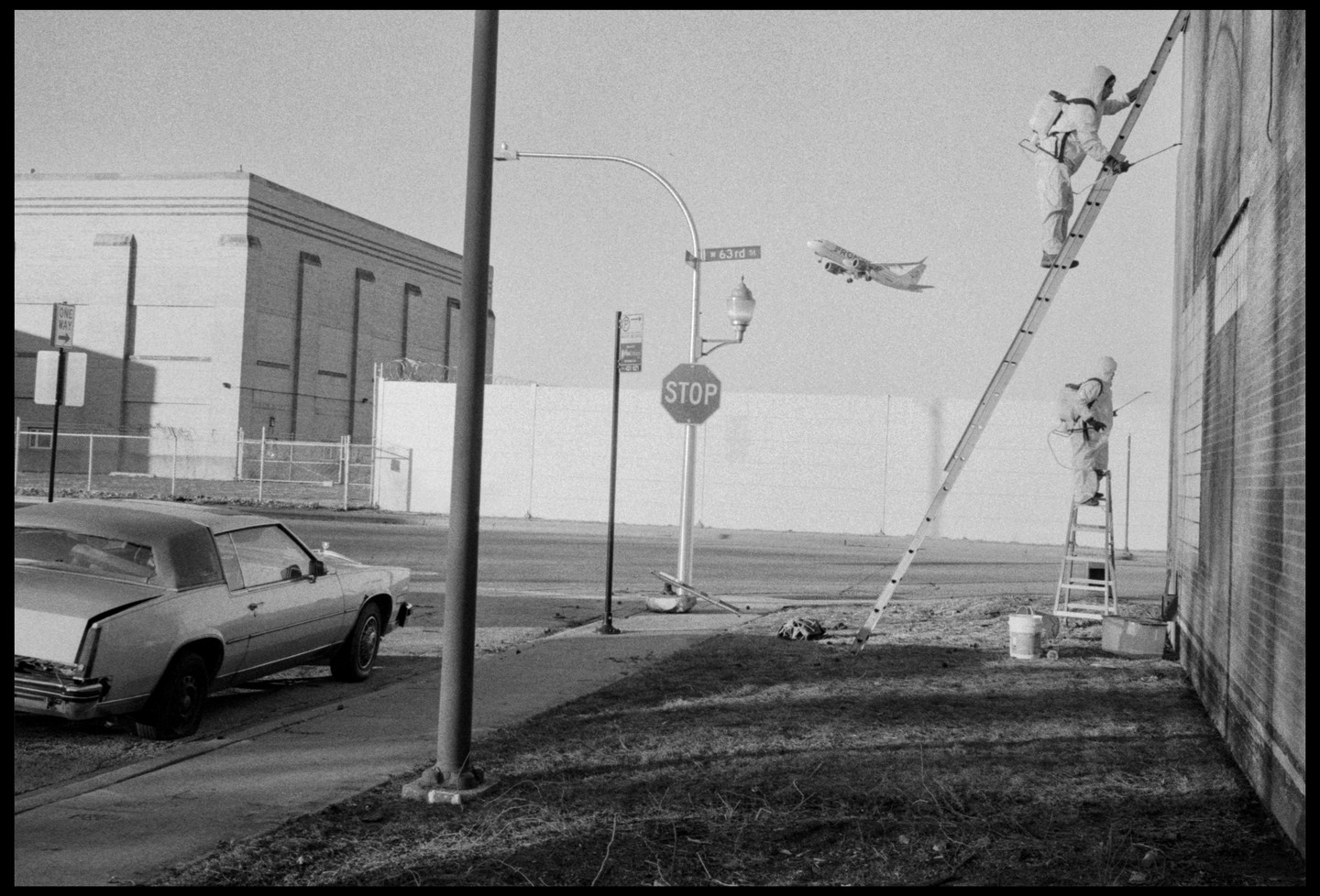 Two people in hazmat suits are on ladders spraying a building as an airplane flies overhead near a stop sign and a parked car.