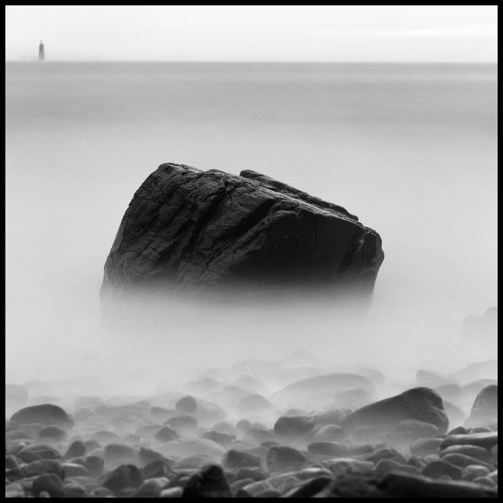 A large boulder sits in the water with smaller rocks around it and a lighthouse in the distance.