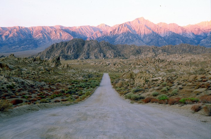 A dirt road leads through a rocky, arid landscape towards mountains illuminated by the setting sun.