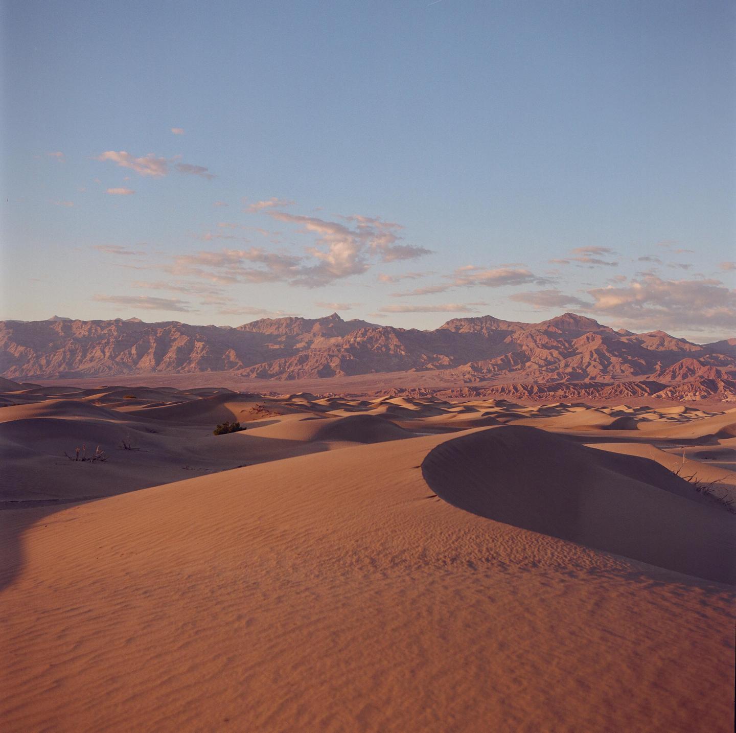 A vast desert landscape features sand dunes in the foreground and mountains in the distance under a partly cloudy sky.