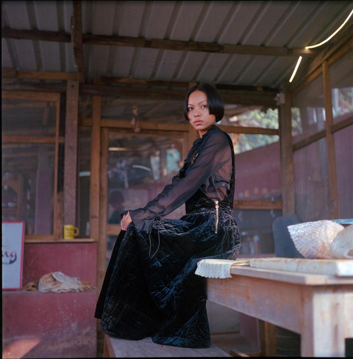 A woman in a black dress sits on a wooden bench in a rustic building.