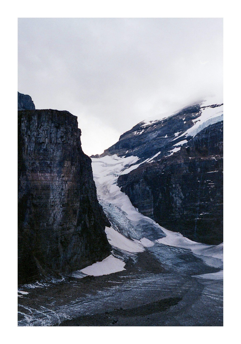 A glacier flows between two dark, rocky mountains under a cloudy sky.