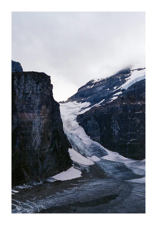 A glacier flows between two dark, rocky mountains under a cloudy sky.