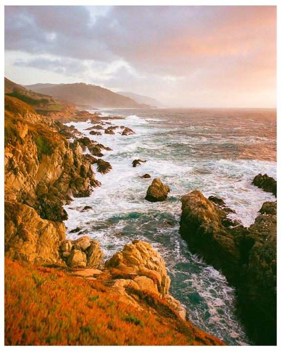 Waves crash against a rocky coastline under a cloudy sky with a pink hue.