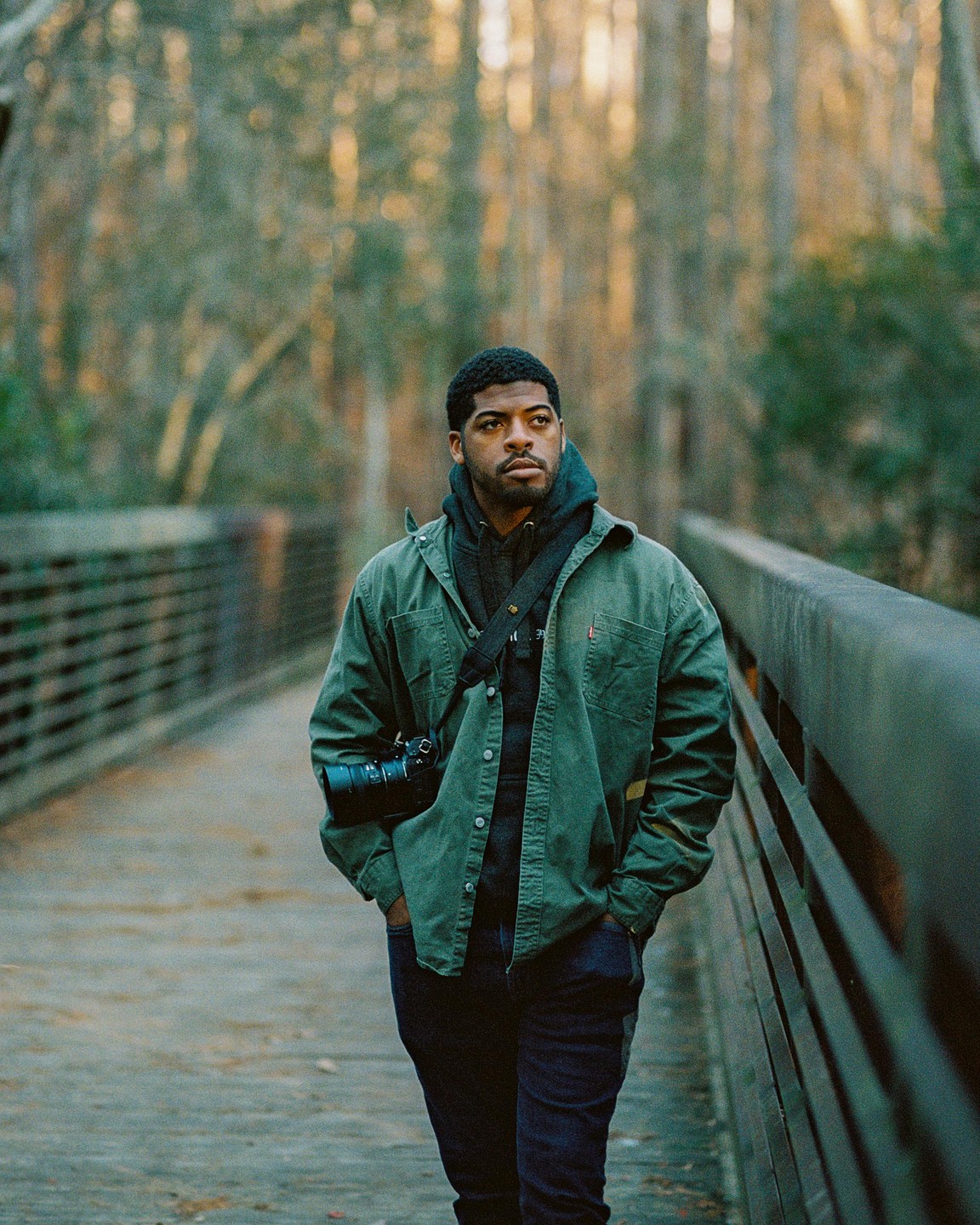 A man with a camera walks on a bridge through a forest.