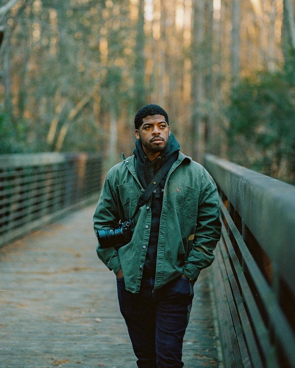 A man with a camera walks on a bridge through a forest.