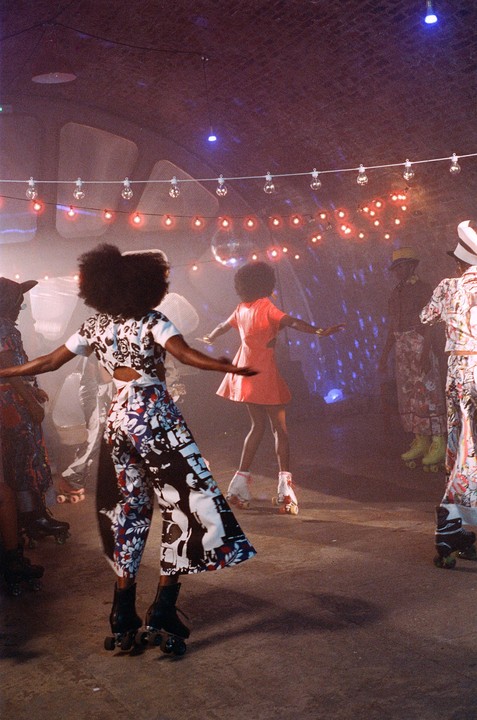 Several people on roller skates are dancing under string lights and a disco ball in a dimly lit room.