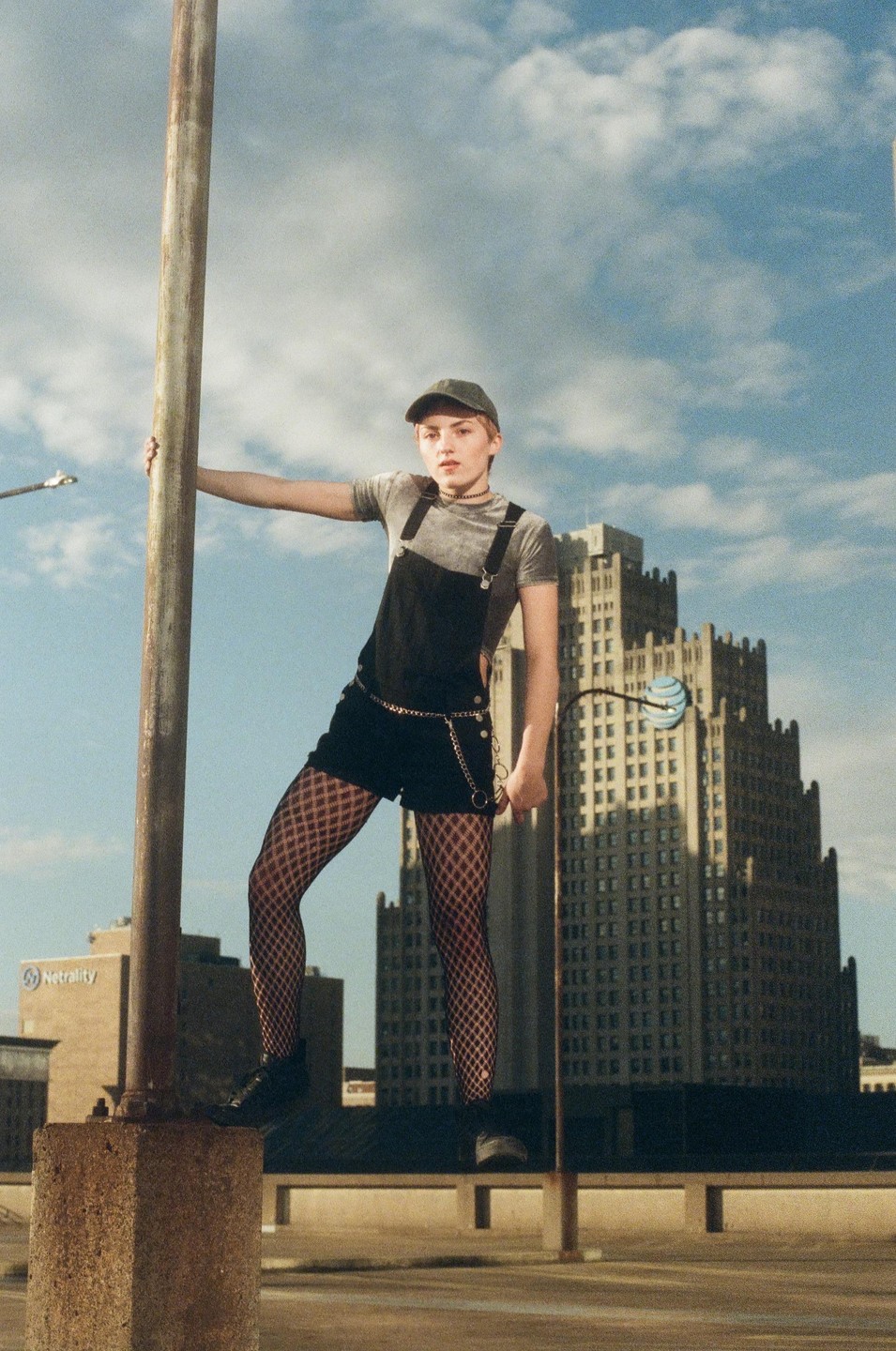 A woman in a baseball cap and overalls poses on a concrete pillar with a city skyline in the background.